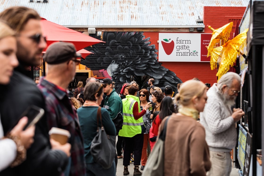 A crowd of people at Farm Gate Market lining up at a stall. in the back ground are yellow umbrellas and a graffiti mural of a black cockatoo.
